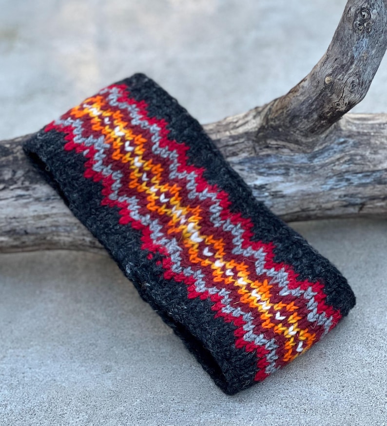 May include: A dark gray knitted headband with a zig-zag pattern in red, orange, yellow, white, and gray. The headband is made of a soft, textured material and is designed to keep the ears warm. The accessory is suitable for cold weather.