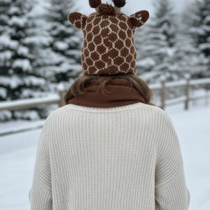 May include: A person wearing a brown and white giraffe-patterned knit hat with small ears and pom-poms. They are also wearing a brown scarf and a cream-colored sweater. The background shows snow-covered trees.