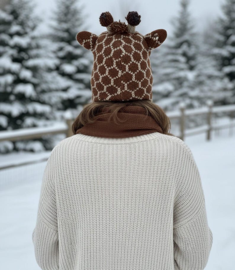 May include: A person wearing a brown and white giraffe-patterned knit hat with small ears and pom-poms. They are also wearing a brown scarf and a cream-colored sweater. The background shows snow-covered trees.
