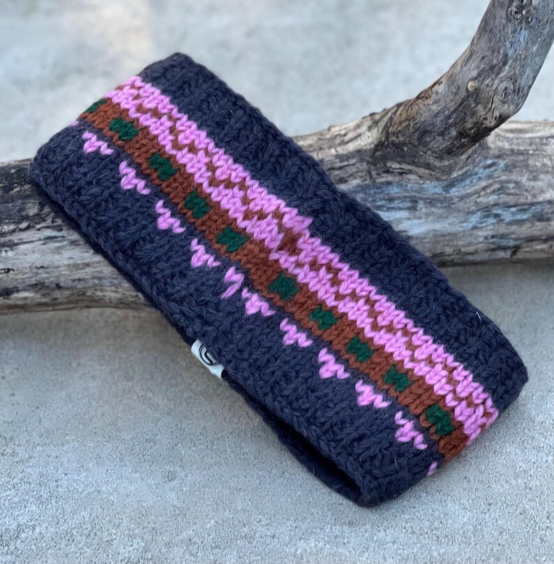 May include: A dark gray knit headband with a geometric pattern in pink, brown, and green. The headband is made of a thick, textured yarn and is designed to keep the ears warm. The accessory is shown on a natural wood background.