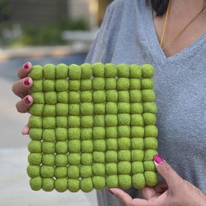 May include: A square, lime green felt trivet composed of numerous small, rounded felt balls. The trivet is held by a person with pink nail polish. The background is blurred, suggesting an outdoor setting.