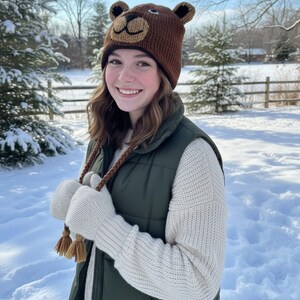 May include: A brown knit bear hat with ear flaps and a bear face design. The hat is worn with a green vest, a cream-colored sweater, and white knit gloves with tassels. The background is a snowy winter scene.