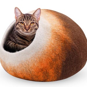 May include: A brown tabby cat peeks out from a cozy, oval-shaped cat bed. The bed is a blend of white, orange, and brown felted wool, creating a warm and inviting space for a feline friend.