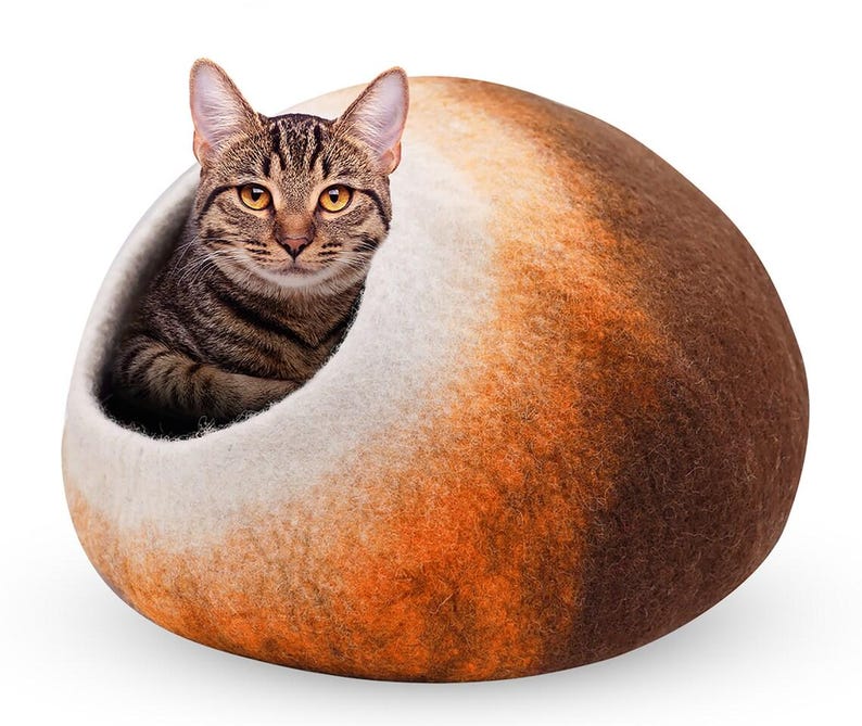 May include: A brown tabby cat peeks out from a cozy, oval-shaped cat bed. The bed is a blend of white, orange, and brown felted wool, creating a warm and inviting space for a feline friend.