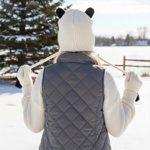 May include: A person wearing a white knit hat with black bear ears, a gray quilted vest, and white gloves with black tassels. The person is standing in a snowy outdoor setting with a tree and a fence in the background.