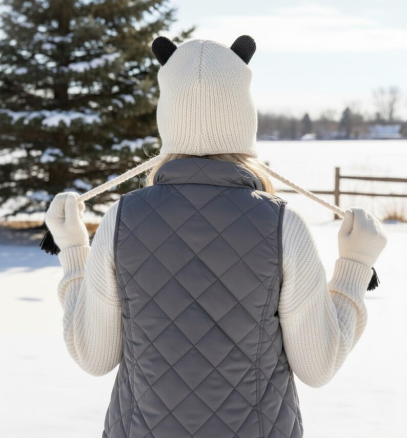 May include: A person wearing a white knit hat with black bear ears, a gray quilted vest, and white gloves with black tassels. The person is standing in a snowy outdoor setting with a tree and a fence in the background.