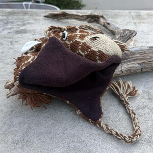 May include: A brown and tan giraffe hat with a fleece-lined face and ear flaps. The hat features a braided tie and white accents, creating a whimsical animal design. The hat is made of knitted material.