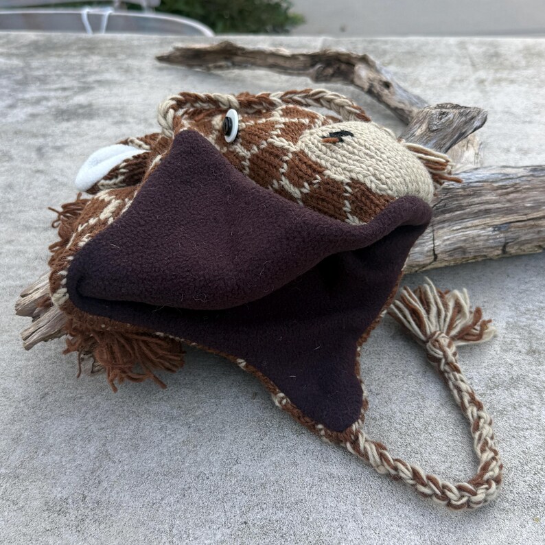 May include: A brown and tan giraffe hat with a fleece-lined face and ear flaps. The hat features a braided tie and white accents, creating a whimsical animal design. The hat is made of knitted material.