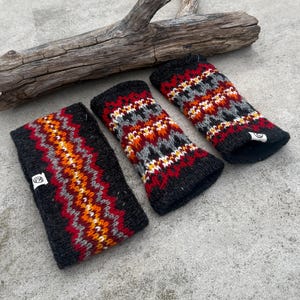 May include: Three black knitted wrist warmers and a headband with a colorful geometric pattern in red, orange, gray, and white. The items are displayed on a concrete surface with a piece of driftwood in the background. Each item has a small white tag.