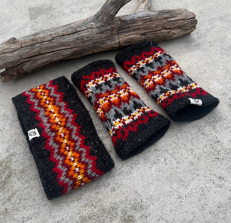 May include: Three black knitted wrist warmers and a headband with a colorful geometric pattern in red, orange, gray, and white. The items are displayed on a concrete surface with a piece of driftwood in the background. Each item has a small white tag.