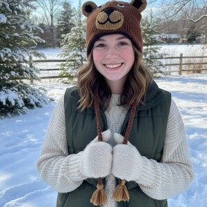 May include: A brown bear-shaped knit hat with ear flaps and a bear face design. The person is wearing a green vest, a cream-colored sweater, and white knit mittens with brown tassels. The background is a snowy winter scene.