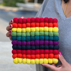 May include: A square, rainbow-colored felt ball trivet. The trivet is made of rows of colorful felt balls in a spectrum of colors, including red, orange, yellow, green, blue, indigo, and violet. The trivet is held by a person.