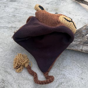May include: A brown knit hat with a lion face design. The hat features a dark brown fleece lining and a braided tail with a tassel. The lion's face is tan with black accents.