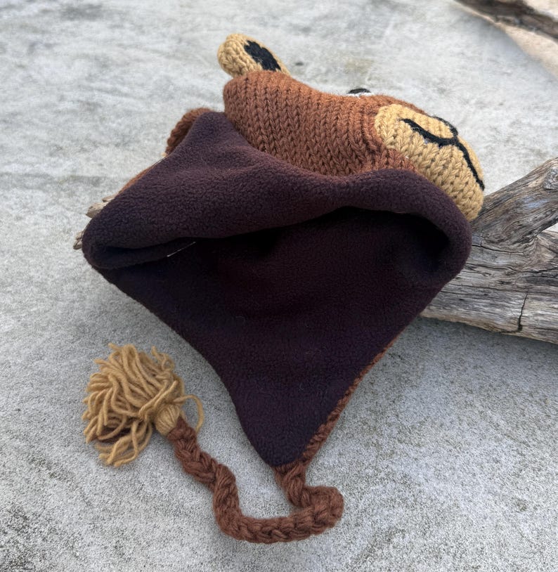 May include: A brown knit hat with a lion face design. The hat features a dark brown fleece lining and a braided tail with a tassel. The lion's face is tan with black accents.