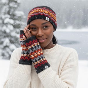 May include: A knitted winter hat and fingerless gloves set. The hat and gloves feature a geometric pattern in red, orange, gray, and black. The woman in the image is wearing a cream-colored sweater.