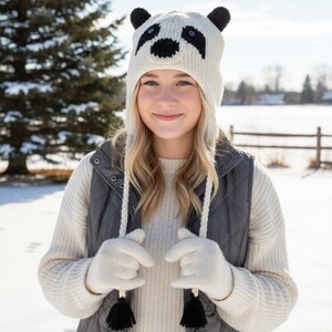 May include: A knitted panda hat with black and white details, featuring ear flaps and a tassel tie. The person is wearing a white sweater, gray vest, and white gloves. The background shows a snowy landscape.