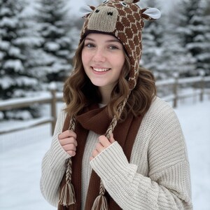 May include: A brown and tan giraffe hat with ear flaps and a matching brown scarf. The hat features a giraffe face with a patterned design. The scarf has braided tassels. The person is wearing a cream-colored sweater.