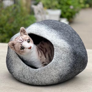 May include: A gray cat bed with a cat-shaped opening. A plush tabby cat with brown and white markings is nestled inside the bed, looking out. The bed is made of a soft, felt-like material, and the background is blurred.