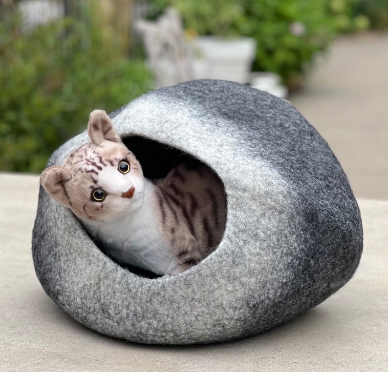 May include: A gray cat bed with a cat-shaped opening. A plush tabby cat with brown and white markings is nestled inside the bed, looking out. The bed is made of a soft, felt-like material, and the background is blurred.
