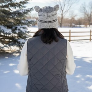 May include: A person wearing a gray quilted vest and a knitted hat with ear-like protrusions. The hat has a striped design. The background shows a snowy landscape with a fence and trees.