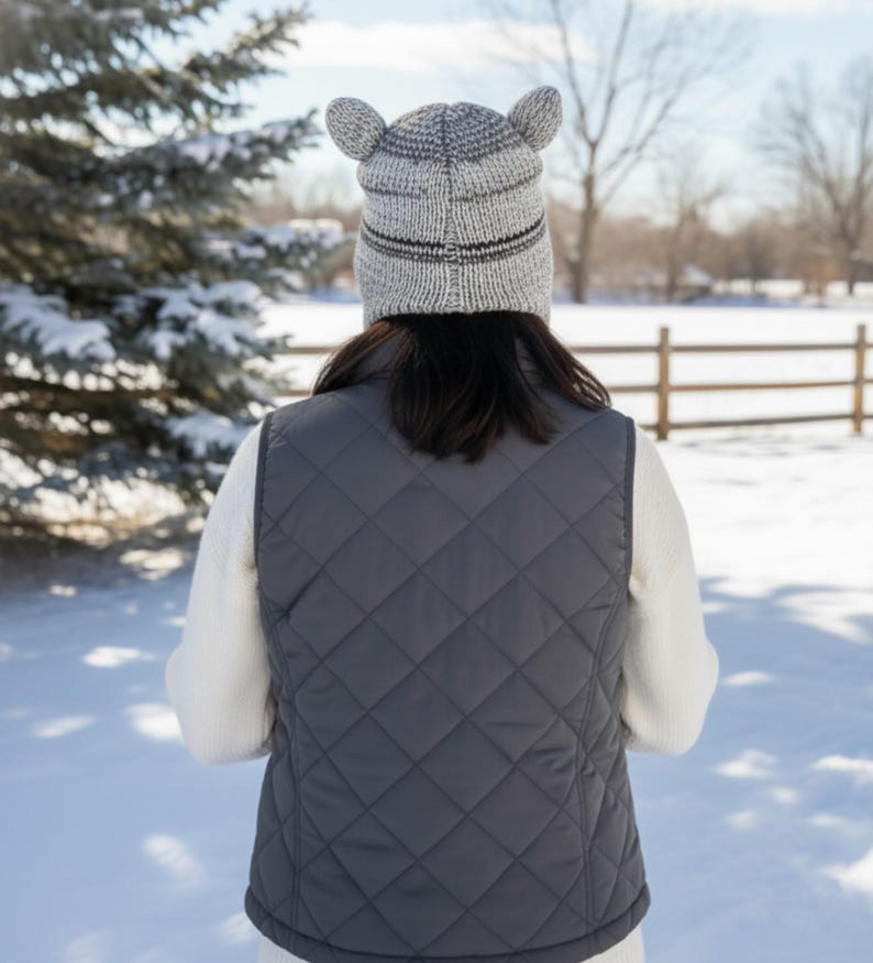 May include: A person wearing a gray quilted vest and a knitted hat with ear-like protrusions. The hat has a striped design. The background shows a snowy landscape with a fence and trees.