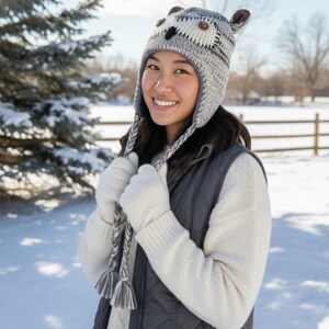 May include: A gray and white knit owl hat with ear flaps and button details. The hat is worn with a white sweater, gray vest, and white gloves. The background shows a snowy winter scene.