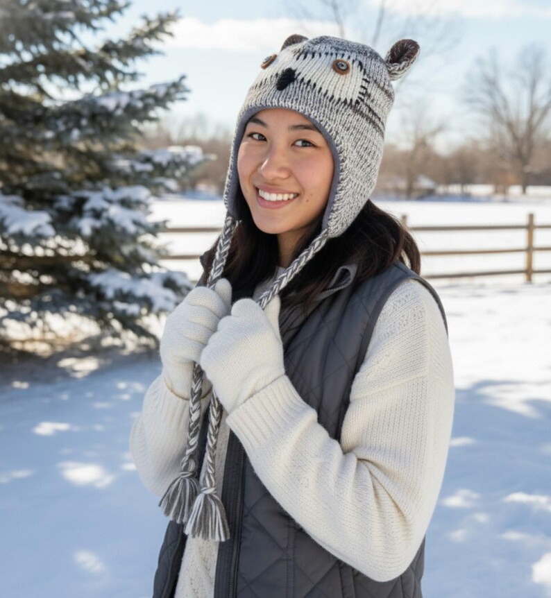 May include: A gray and white knit owl hat with ear flaps and button details. The hat is worn with a white sweater, gray vest, and white gloves. The background shows a snowy winter scene.