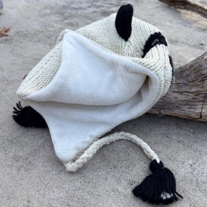 May include: A white and black knit hat with a fleece lining. The hat features black ear accents and a braided white cord with a black tassel. The hat is designed to keep the head warm.