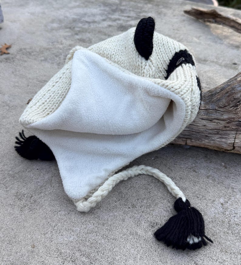 May include: A white and black knit hat with a fleece lining. The hat features black ear accents and a braided white cord with a black tassel. The hat is designed to keep the head warm.