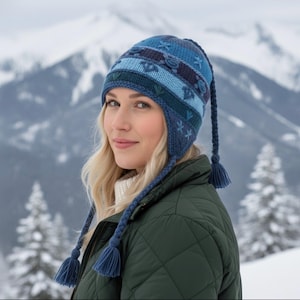May include: A blue knit hat with ear flaps and long braided ties ending in tassels. The hat features a pattern of floral and geometric designs in shades of blue. The background shows snow-covered mountains.