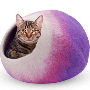 May include: A tabby cat peeks out from a felted cat bed. The bed is a rounded shape with a gradient of colors, transitioning from white to pink and purple. The cat has brown and black stripes and yellow eyes.