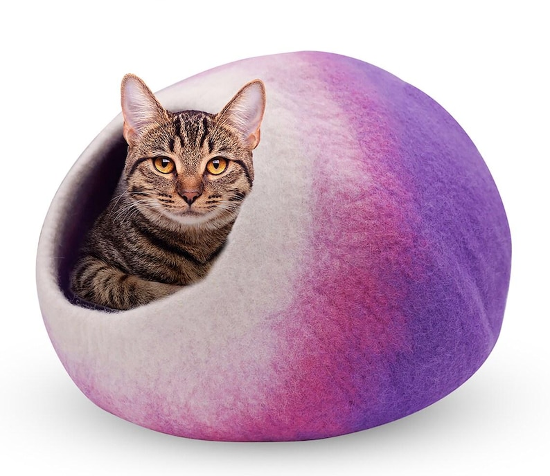 May include: A tabby cat peeks out from a felted cat bed. The bed is a rounded shape with a gradient of colors, transitioning from white to pink and purple. The cat has brown and black stripes and yellow eyes.