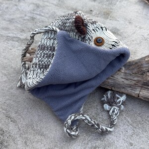 May include: A handmade knit hat with a gray and white pattern, featuring a blue fleece lining. The hat has a button eye, brown ear accents, and braided ties with a pom-pom. The hat is designed to resemble an animal.
