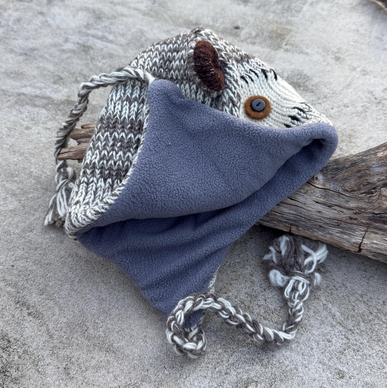 May include: A handmade knit hat with a gray and white pattern, featuring a blue fleece lining. The hat has a button eye, brown ear accents, and braided ties with a pom-pom. The hat is designed to resemble an animal.