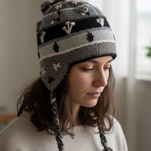 May include: A knitted winter hat with ear flaps, featuring a pattern of mushrooms, leaves, and stars. The hat is primarily gray, black, and white, with braided tassels. The hat is designed to keep the wearer warm during cold weather.
