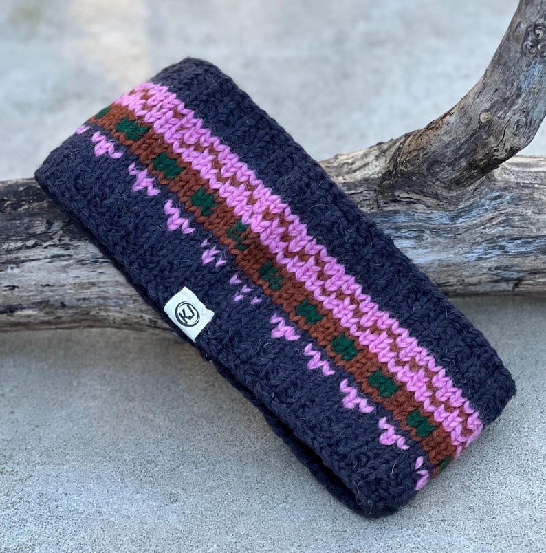 May include: A dark blue knitted headband with a geometric pattern in pink, brown, and green. The headband has a small white tag with a circular logo. The accessory is designed to keep the ears warm.