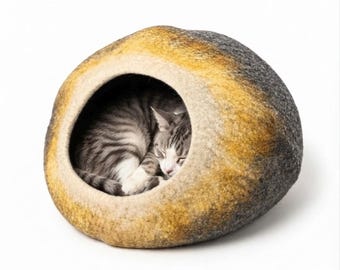 May include: A rounded, felted wool cat cave with a textured surface in shades of charcoal gray, mustard yellow, and cream. A gray and white tabby cat is curled up inside the circular opening of the soft, stone-shaped pet bed.