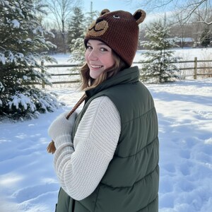 May include: A brown knit bear hat with ear flaps and a bear face design. The hat is worn with a green puffer vest, a cream-colored sweater, and white gloves. The background is a snowy winter scene.