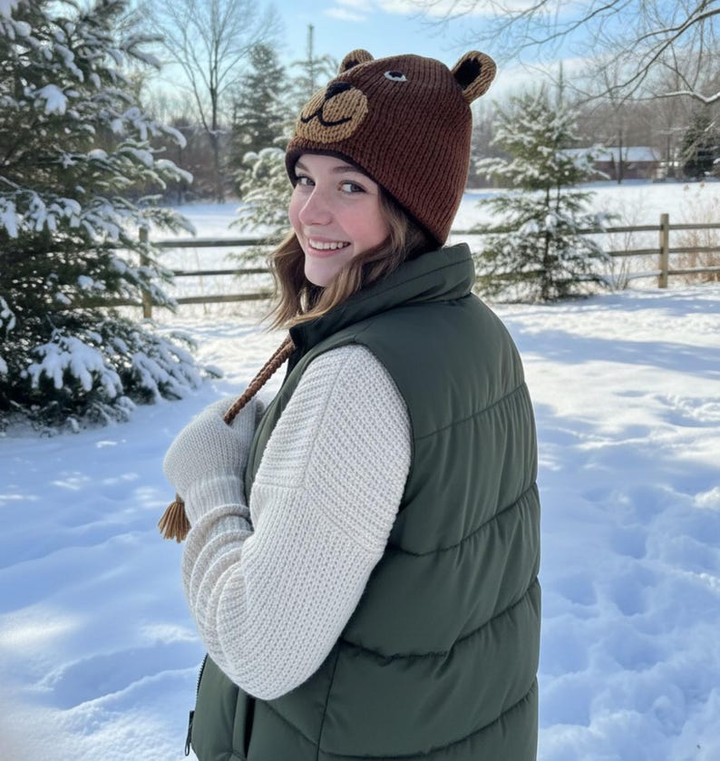May include: A brown knit bear hat with ear flaps and a bear face design. The hat is worn with a green puffer vest, a cream-colored sweater, and white gloves. The background is a snowy winter scene.