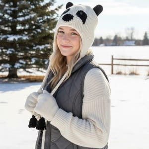 May include: A cream-colored knit panda hat with black eyes and ears. The hat has a matching cream-colored chin strap with black tassels. The person is wearing a gray quilted vest and cream-colored gloves.