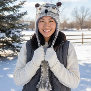 May include: A knitted owl hat with ear flaps and braided ties. The hat features brown ear accents, button eyes, and a black beak. The person in the image is wearing a white sweater, gray vest, and white gloves, standing in a snowy outdoor setting.