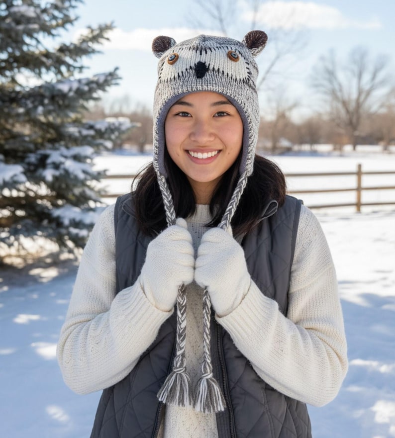 May include: A knitted owl hat with ear flaps and braided ties. The hat features brown ear accents, button eyes, and a black beak. The person in the image is wearing a white sweater, gray vest, and white gloves, standing in a snowy outdoor setting.