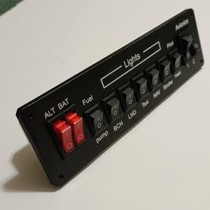 May include: A black control panel with multiple switches and the text "Lights." The panel includes switches labeled "ALT BAT," "Fuel," "pump," "BCN," "LND," "Taxi," "NAV," "Strobe," "Pilot," and "heat." Two red switches are visible.