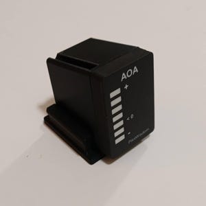 May include: A black rectangular electronic device with a vertical display featuring white segments, the text AOA, a plus sign, a zero, a minus sign, and the brand name PilotWisdom printed in white on the front face of the housing.