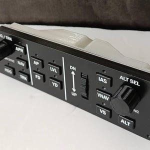 May include: Black control panel with white lettering and various buttons and knobs. The panel includes labels such as HDG/TRK, APR, AP, LVL, IAS, and ALT SEL. A gray plastic housing is attached to the top of the panel.