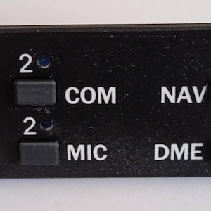 May include: Black control panel with multiple buttons and labels. The panel has buttons labeled COM, NAV, MIC, DME, and ADF. It also has indicator lights and labels like O, M, and I. The panel is rectangular with screw holes on the corners.