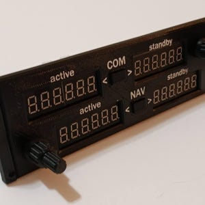 May include: A black electronic panel with multiple digital displays and knobs. The panel features several 7-segment displays showing numerical readouts, labeled 'active' and 'standby'. The panel also has labels for 'COM' and 'NAV'.