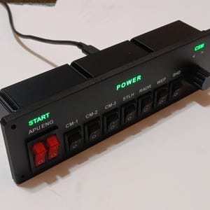 May include: A black rectangular control panel featuring two red toggle switches labeled START APU ENG, followed by six black rocker switches labeled CM-1, CM-2, CM-3, STLH, RADR, WEP, and SHD. The panel includes green backlit text for POWER and CSM, with a rotary knob on the right.