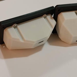 May include: Two white, geometric-shaped electronic devices with black frames. Each device has a small port and is attached with screws. The devices are on a white surface.