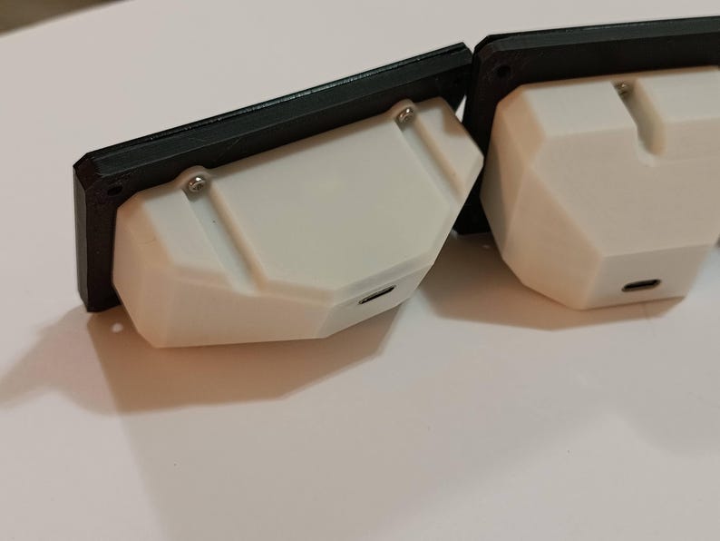 May include: Two white, geometric-shaped electronic devices with black frames. Each device has a small port and is attached with screws. The devices are on a white surface.